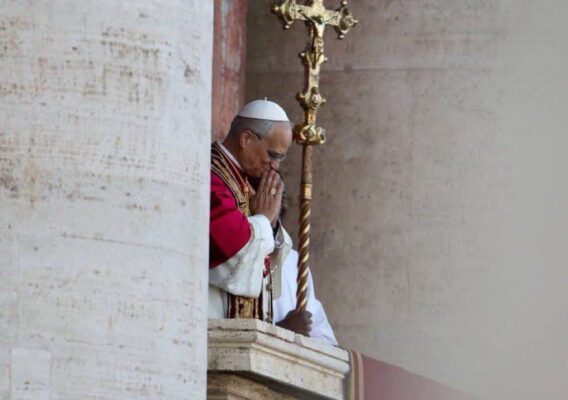 Pope Francis praying from a balcony at the Vatican, dressed in papal robes, with a golden crucifix in the background. The image captures a moment of reflection and spirituality during a public appearance.