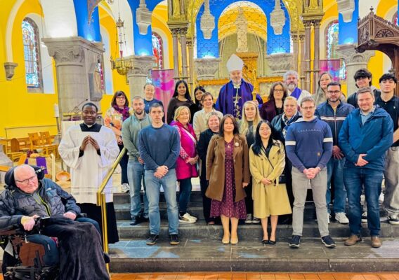 Group photo of a diverse congregation in a church setting, featuring a bishop in ceremonial attire, clergy members, and attendees of various ages. The vibrant interior includes stained glass windows and colorful decorations, highlighting a community gathering or religious event.