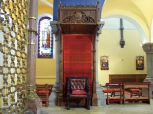Interior view of a church featuring a beautifully carved wooden throne with red upholstery, surrounded by decorative stained glass windows and wooden benches. The warm yellow walls and stone flooring enhance the serene atmosphere of the sacred space.