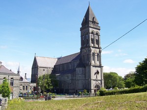 Historic stone church with a tall steeple and intricate architecture, surrounded by greenery under a clear blue sky.
